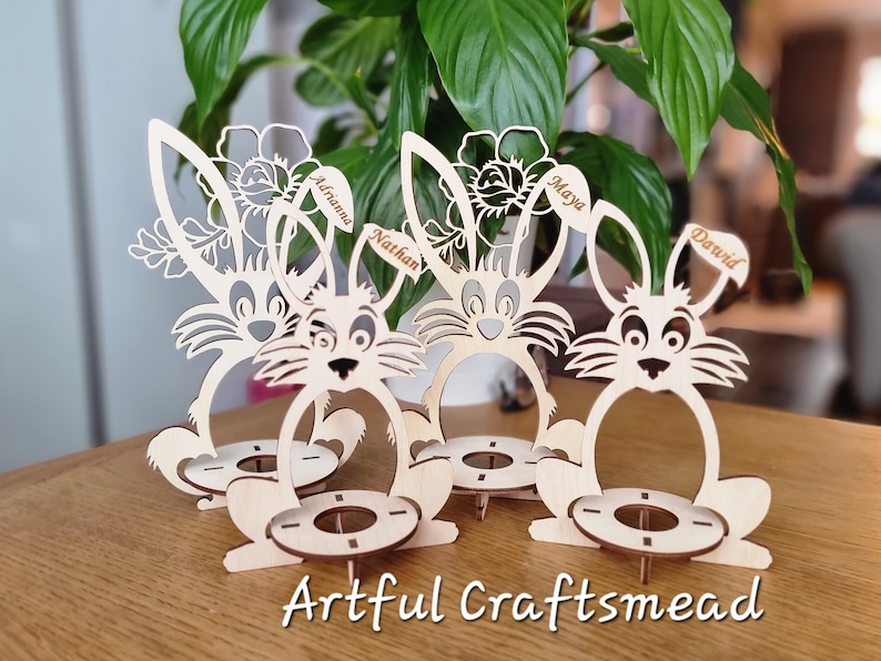 May include: Four wooden bunny egg holders with personalized names, "Adriana", "Nathan", "Maya", and "David",  are arranged on a wooden surface. Each bunny has a flower design and a space for an egg.