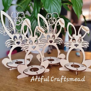 May include: Four wooden bunny egg holders with personalized names:  'Adrian', 'Nathan', 'Maya', and 'David'. Each holder features a bunny design with a flower and a small circular space for an egg.