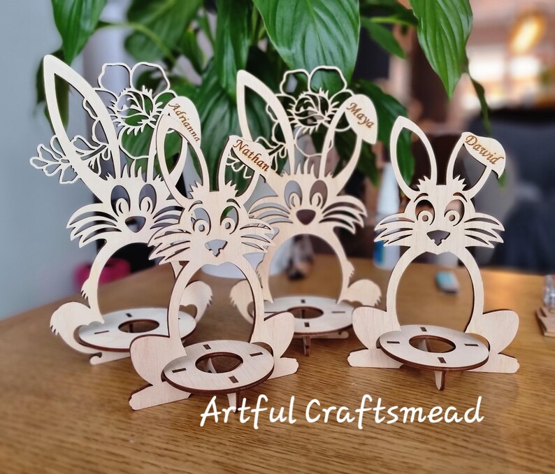 May include: Four wooden bunny egg holders with personalized names:  'Adrian', 'Nathan', 'Maya', and 'David'. Each holder features a bunny design with a flower and a small circular space for an egg.