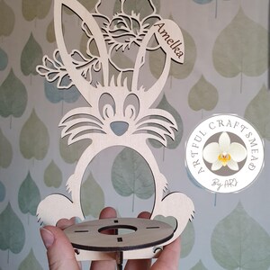 May include: Wooden bunny-shaped Easter egg holder with a floral design and the name "Amelia" engraved on the top. The holder has a round base with a hole in the center for an egg. The holder is made by "Artful Crafts Mead" and is a light brown color.