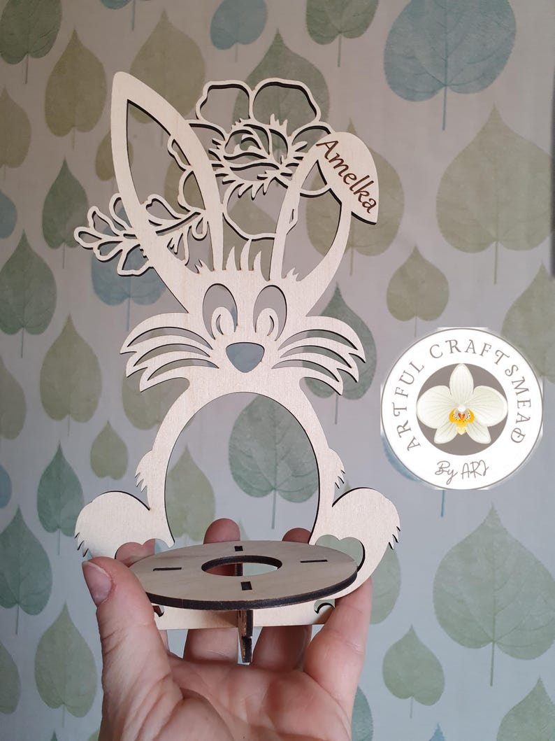 May include: Wooden bunny-shaped Easter egg holder with a floral design and the name "Amelia" engraved on the top. The holder has a round base with a hole in the center for an egg. The holder is made by "Artful Crafts Mead" and is a light brown color.