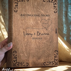 May include: A rectangular wooden book cover with a dark brown finish, featuring laser-engraved corner flourishes, the text AN ONGOING STORY, a double heart graphic, the names Vinny & Brenna, and the date 09/24/2021 centred on the front panel.