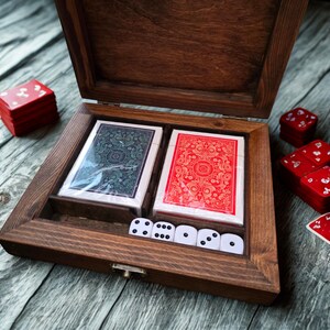 May include: A wooden game box with a hinged lid, containing two decks of playing cards, dice, and small red boxes. One deck is red, the other is blue. The dice are white with black dots. The box is made of dark wood.