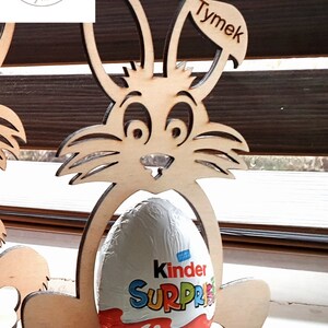 May include: Wooden bunny rabbit Easter egg holder with a Kinder Surprise egg inside. The bunny is holding the egg in its paws and has a name tag that says "Tymek".