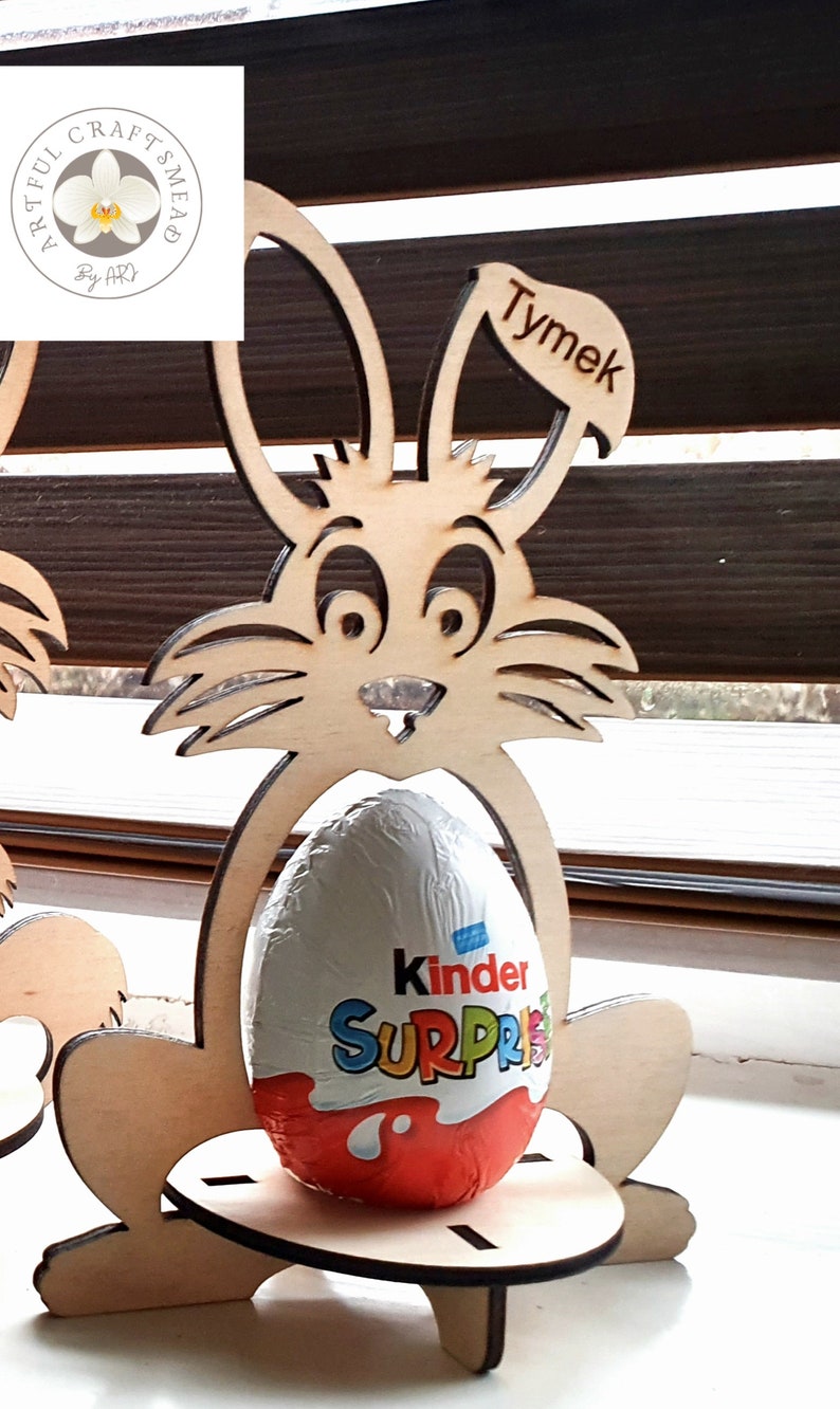 May include: Wooden bunny rabbit Easter egg holder with a Kinder Surprise egg inside. The bunny is holding the egg in its paws and has a name tag that says "Tymek".
