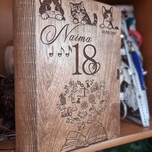 May include: A rectangular wooden box with a laser-engraved design featuring three kittens at the top, the name Naima, the number 18, the date 18/09/2007, musical notes, and a collection of academic icons emerging from an open book at the bottom.