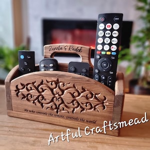 May include: Wooden remote control holder with a carved design and the text "Zaneta & Radek" and "He who controls the remote, controls the world". The holder has four slots for remotes and is made of dark brown wood.