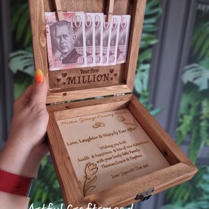 May include: A wooden keepsake box with a hinged lid featuring two small clips holding five ten-pound notes. The interior lid is engraved with "Your first MILLION." The base features a personalised message: "Horton Grange Country House, Love, Laughter & Happily Ever After! Wishing you both health & happiness & lots of new adventures with your lovely little family: Thomas, Gracie & Daphne. Love Jayne & Clive xxx."