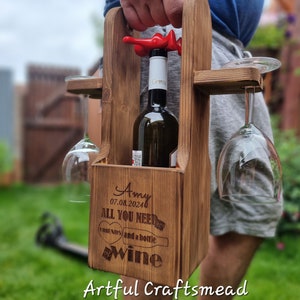 May include: A wooden wine carrier with a bottle of wine and two glasses. The carrier has a handle and is engraved with the text "Amy 07.08.2024 ALL YOU NEED IS FRIENDS and a bottle of WINE".