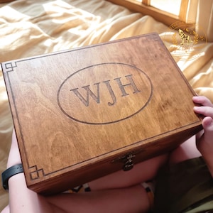 May include: A rectangular wooden box with a dark brown finish, featuring a central oval engraving of the letters WJH and geometric corner accents. The box includes a metal latch on the front edge and a smooth, polished wood grain surface.