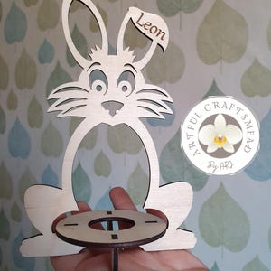 May include: Wooden bunny-shaped Easter egg holder with a round base and a name tag that says "Leon". The holder is made by Artful Craftsmead.