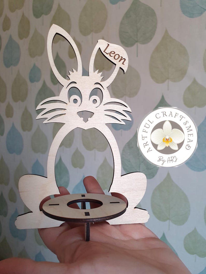 May include: Wooden bunny-shaped Easter egg holder with a round base and a name tag that says "Leon". The holder is made by Artful Craftsmead.