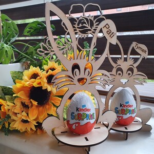 May include: Two wooden bunny-shaped Easter egg holders with the names "Pati" and "Tymek" on them. Each holder has a Kinder Surprise egg inside.