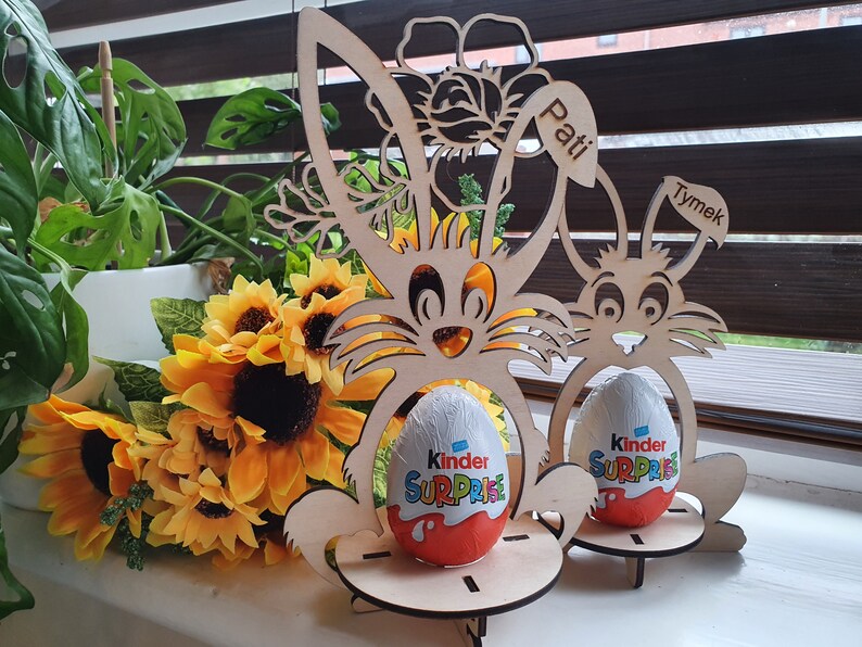 May include: Two wooden bunny-shaped Easter egg holders with the names "Pati" and "Tymek" on them. Each holder has a Kinder Surprise egg inside.
