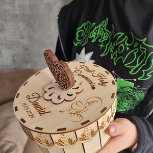 May include: A round, light-colored wooden box with a decorative heart-shaped handle. The lid is engraved with the words "First Holy Communion", the name "David", and the date "18.05.2024". The box features a floral design and chalice illustrations.