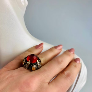 Peut inclure: Une bague en or avec une grande pierre rouge entourée de petites pierres claires. La bague présente un design détaillé avec des sculptures complexes.