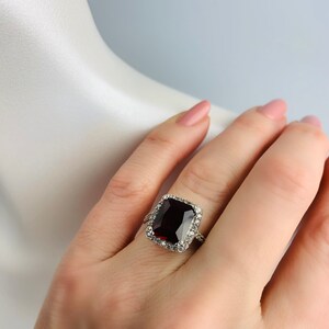 May include: A silver ring with a large, dark red, square-shaped gemstone surrounded by a halo of smaller, clear gemstones.