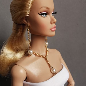 Könnte beinhalten: Eine Puppe mit blonden Haaren, die ein weißes, trägerloses Oberteil und eine goldene Kettenkette mit weißen Perlenakzenten trägt. Die Puppe trägt auch goldene Ohrringe mit weißen Perlenakzenten.