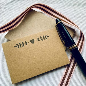 Peut inclure: Une carte brune avec un simple motif de cœur et de feuilles. La carte est posée sur une surface blanche avec un ruban rayé rose et brun et un stylo noir.
