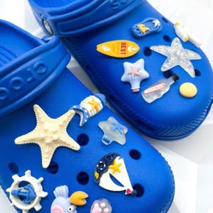 May include: Bright blue clogs decorated with various beach-themed charms, including starfish, a surfboard, and a sailboat. The clogs have a textured surface and are designed for casual wear. The text "Style B" is visible.