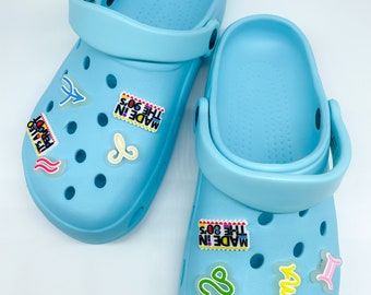 May include: Two light blue clogs with attached straps. Each clog has several colorful decorative charms, including zodiac symbols and text that reads "MADE IN THE 90'S" and "IT'S ALSO PERIODT". The clogs are made of a rubbery material.