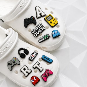 May include: White clogs decorated with colorful gaming-themed charms. The charms include game controllers, pixelated ghosts, letters, and a handheld game console. One clog has the text "GAME OVER" and a battery icon.