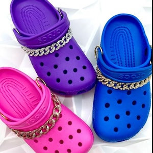May include: Three colorful Crocs shoes: purple, blue, and pink. Each shoe has a decorative chain around the top. The shoes are on a white surface.
