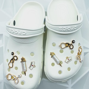 May include: A pair of white Crocs shoes adorned with medical-themed charms. The charms include stethoscopes, syringes, and a nurse's cap, all in gold and white. The shoes are set against a white background.