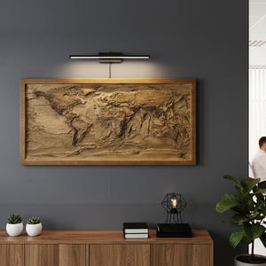 Könnte beinhalten: Eine große, gerahmte Weltkarte aus Holz mit detailliertem Relief der Kontinente. Die Karte ist an einer dunkelgrauen Wand befestigt und wird von einer schwarzen Leuchte beleuchtet. Ein Holzschrank und Pflanzen befinden sich im Vordergrund.