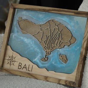 Könnte beinhalten: Holzgerahmtes Wandbild mit einer geschichteten Karte von Bali. Die Karte besteht aus hellbraunem Holz, vor einem blauen Harzhintergrund und ist in dunklem Holz gerahmt. Das Wort "BALI" ist unten eingraviert.
