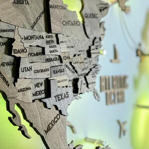 Op de afbeelding: Een meerlaagse houten wandkaart van Noord-Amerika met in laser gegraveerde namen van staten en provincies zoals Californi&euml;, Texas en Ontario. De kaart is bevestigd met zilverkleurige hardware tegen een fel limoengroene achtergrond.
