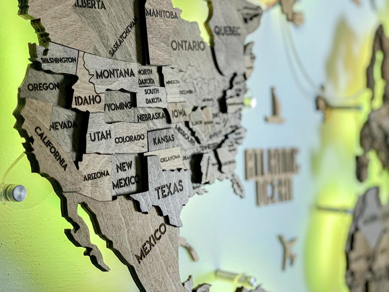 Op de afbeelding: Een meerlaagse houten wandkaart van Noord-Amerika met in laser gegraveerde namen van staten en provincies zoals Californi&euml;, Texas en Ontario. De kaart is bevestigd met zilverkleurige hardware tegen een fel limoengroene achtergrond.