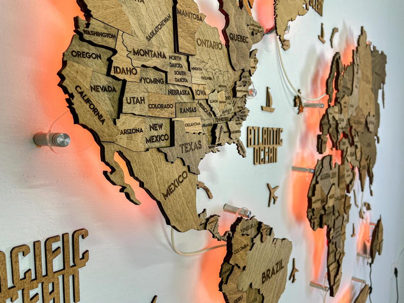 Op de afbeelding: Een meerlaagse houten wereldkaart als wanddecoratie met lasergesneden continenten en landen met gegraveerde labels. De kaart is gemonteerd met afstandhouders die een achtergrondverlichtingseffect cre&euml;ren met warm oranje licht.