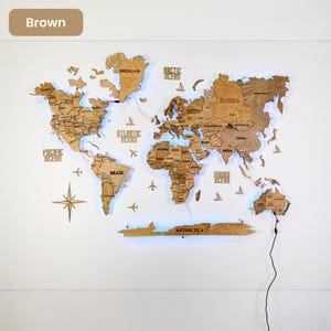 Op de afbeelding: Een bruine houten wereldkaart als wanddecoratie met lasergesneden continenten, landen en oceanen. De kaart bevat labels voor regio's, een windroos, vliegtuigiconen en scheepssilhouetten, voorzien van ge&iuml;ntegreerde blauwe LED-achtergrondverlichting.