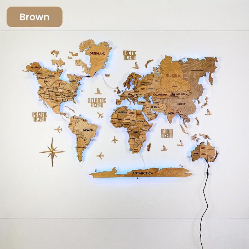 Op de afbeelding: Een bruine houten wereldkaart als wanddecoratie met lasergesneden continenten, landen en oceanen. De kaart bevat labels voor regio's, een windroos, vliegtuigiconen en scheepssilhouetten, voorzien van ge&iuml;ntegreerde blauwe LED-achtergrondverlichting.