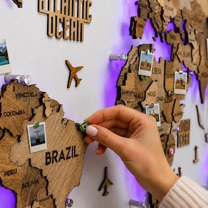 Può includere: Una mappa del mondo in legno da parete con continenti tagliati al laser e nomi dei paesi incisi. La mappa include piccole icone di aerei in legno, distanziatori in metallo, retroilluminazione a LED viola e diverse foto appuntate.