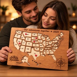 May include: A rectangular wooden map of the United States featuring laser-cut state borders, the text "Our adventures together 2021", and a compass rose. Small red heart markers are placed on specific states, and the word "Adventure" is engraved.