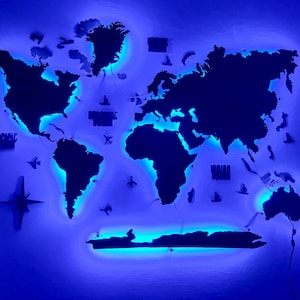Op de afbeelding: Een donkergekleurde wereldkaart als wanddecoratie met een levendige blauwe LED-achtergrondverlichting die de continenten verlicht. De kaart bevat kleine decoratieve iconen van vliegtuigen en schepen, samen met een kompasroos aan de linkerzijde.