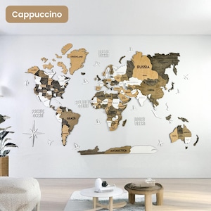 Op de afbeelding: Een meerlaagse houten wereldkaart als wanddecoratie in tinten bruin, beige en wit. De kaart toont gelabelde continenten, landen en oceanen, geaccentueerd met kleine witte houten vliegtuig- en kompasiconen op een witte muur.