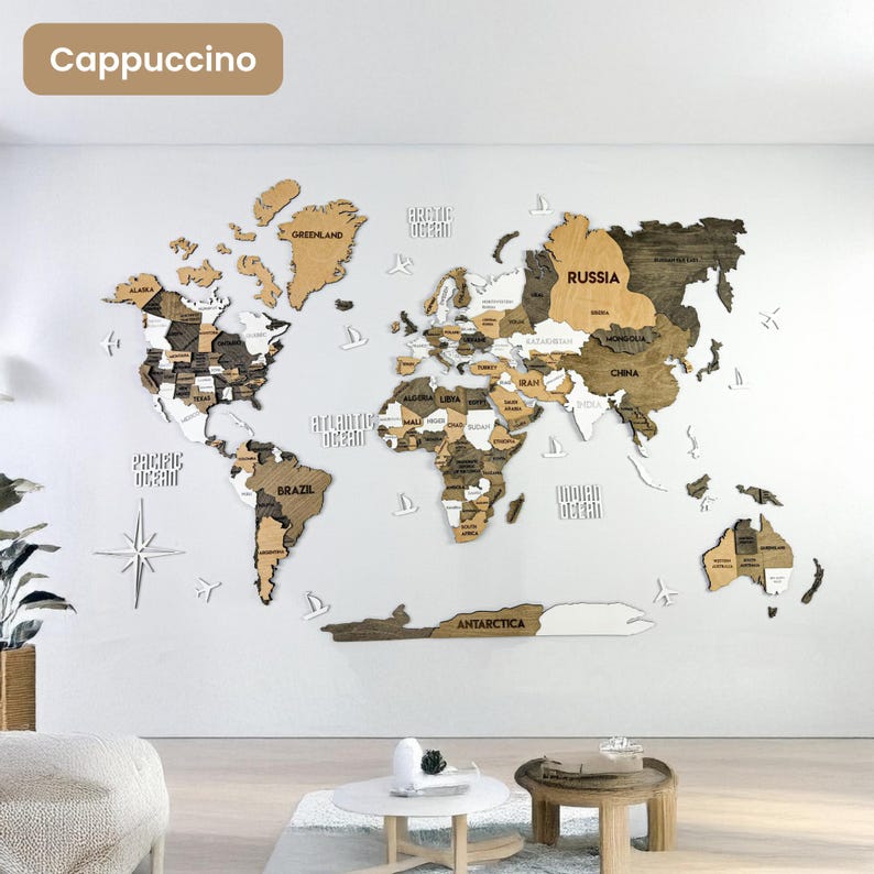 Op de afbeelding: Een meerlaagse houten wereldkaart als wanddecoratie in tinten bruin, beige en wit. De kaart toont gelabelde continenten, landen en oceanen, geaccentueerd met kleine witte houten vliegtuig- en kompasiconen op een witte muur.