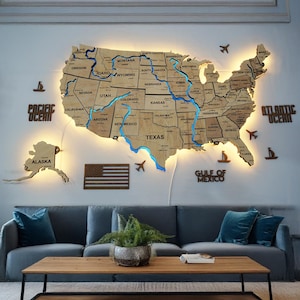 May include: A wooden map of the United States with illuminated blue rivers and state outlines. The map includes Alaska and is surrounded by wooden cutouts of airplanes, boats, and the words "PACIFIC OCEAN", "ATLANTIC OCEAN", and "GULF OF MEXICO".