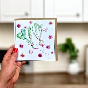 May include: A square greeting card with a watercolour illustration of radishes. The card features green leaves, pink and white bulbs, and a gold border. The card is held against a neutral background.