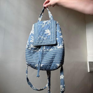 Puede incluir: Mochila acolchada azul con un estampado floral y a rayas. La mochila tiene cierre de solapa, cordón y correas ajustables. La bolsa está sujeta por una mano.