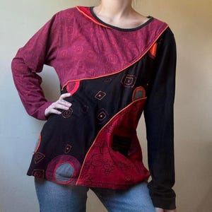 Puede incluir: Una blusa de manga larga con diseño patchwork. La blusa presenta una sección superior burdeos con un patrón circular, un panel central negro y una sección inferior roja. El diseño está delineado con ribetes naranjas. La manga derecha es negra.
