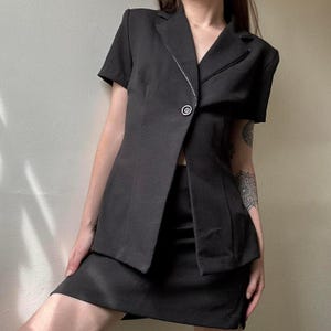 May include: A black short-sleeved blazer and matching skirt set. The blazer features a single button closure and a contrasting trim. The skirt is a mini skirt. The outfit is a classic, tailored style.
