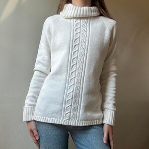 May include: Cream-colored turtleneck sweater with a central cable knit design. The long-sleeved sweater features ribbed cuffs and hem, offering a classic and cozy look. The sweater is made of a soft, textured material.