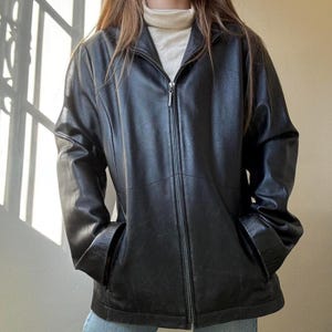 May include: A black leather jacket featuring a full-length front zipper closure, a high collar, and long sleeves. The jacket has a smooth texture, visible seam detailing across the chest and torso, and two side pockets at the waistline.