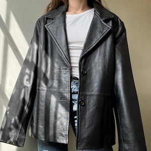 May include: A black leather blazer with a notched collar and two-button closure. The jacket has a classic design and is worn over a white ribbed tank top and blue jeans.