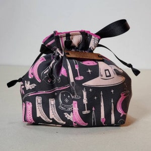 May include: A black drawstring bag with a pink and white print of various witchy items, including boots, candles, a cauldron, and a witch's hat. The bag has a black drawstring and a wooden tag that says "Witchy Bag".