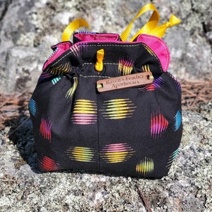 Puede incluir: Una bolsa de cordón negro con un patrón geométrico colorido. La bolsa tiene una etiqueta de madera que dice "Raven's Feather Apothecary".